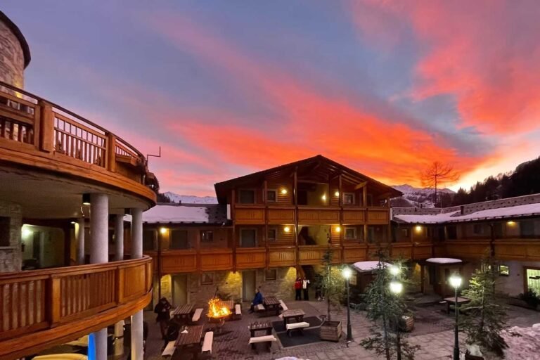 Cortile dell'Hotel Boscone al tramonto, con edifici in legno e pietra e un braciere acceso